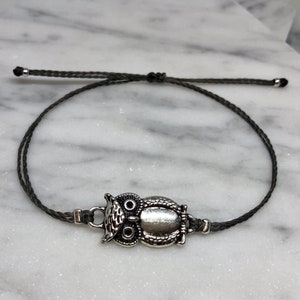 Puede incluir: Pulsera con dije de búho plateado y cordón gris oscuro. El dije de búho presenta detalles de plumas y ojos grabados. La pulsera es ajustable con nudos corredizos.