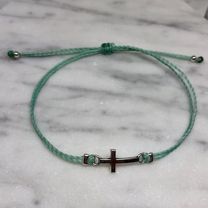 May include: A light green adjustable bracelet featuring a silver cross charm. The bracelet is made of a thin, woven cord with a sliding knot closure. Small silver beads are at the ends of the cord.