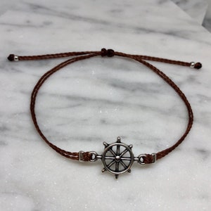 Peut inclure: Un bracelet en cordon marron avec un charm en forme de gouvernail argenté. Le bracelet a des nœuds coulissants réglables et de petites perles argentées aux extrémités. Le bracelet sur le thème nautique est présenté sur une surface en marbre blanc.