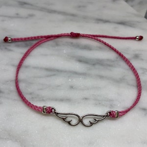 May include: A pink string bracelet featuring a silver-colored wing charm. The bracelet is adjustable with a sliding knot closure and small silver beads at the ends. The wing charm is a detailed, symmetrical design.