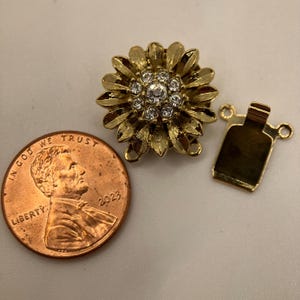 Puede incluir: Un cierre de joyería dorado con diseño de flor, con brillantes transparentes en el centro. Junto a él, una pequeña pieza rectangular dorada con un lazo. Una moneda de cobre de 2023 también está en la imagen.