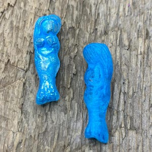 Puede incluir: Dos pequeñas figuras de sirenas azul brillante con detalles. Las sirenas están sobre una superficie de madera desgastada. Las figuras tienen cabello suelto y colas de pez.