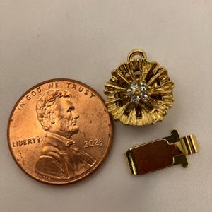 Puede incluir: Un dije de joyería dorado con un diseño floral y pequeñas piedras transparentes en el centro. Un cierre dorado y una moneda de un centavo están al lado del dije. La moneda está fechada en 2023.