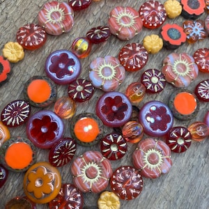 May include: Close-up of various glass beads in shades of red, orange, and gold. The beads feature floral and geometric designs, with some having a striped or speckled appearance. The beads are arranged on a wooden surface.