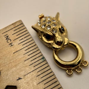 Puede incluir: Un cierre metálico dorado con forma de cabeza de jaguar, adornado con pedrería transparente y ojos negros. El cierre tiene un anillo y tres pequeños bucles. Una regla muestra que el cierre mide aproximadamente 2,5 cm de largo.