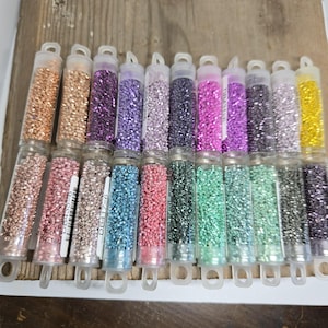 Puede incluir: Una colección de pequeños tubos de plástico transparente llenos de cuentas de colores. Las cuentas vienen en una variedad de colores, incluyendo dorado, morado, rosa, azul, verde y negro. Cada tubo tiene una etiqueta blanca con un código. Los tubos están dispuestos en filas.