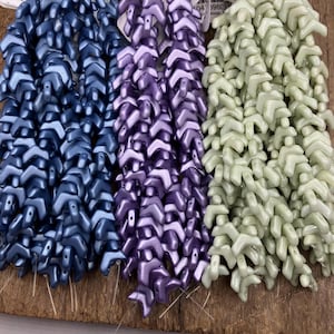 May include: Close-up of three strands of chevron-shaped beads in shades of blue, purple, and green. The beads are strung together, creating a textured and layered effect. The beads are likely for jewelry making.
