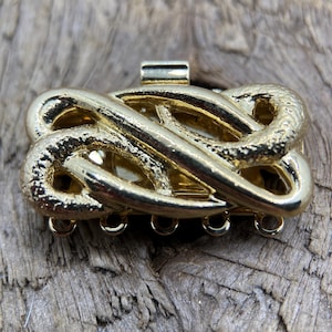 Puede incluir: Un cierre de joyería dorado con un diseño intrincado e interconectado. El cierre tiene forma rectangular con una superficie texturizada y múltiples bucles para la fijación. La estética general es vintage y ornamentada.