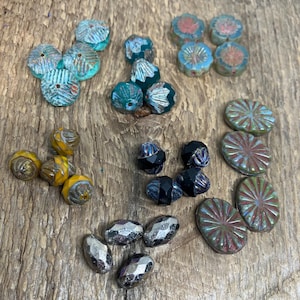 Puede incluir: Una colección de varias cuentas coloridas en diferentes formas y tamaños. Las cuentas incluyen turquesa, verde azulado, amarillo, negro y plata, con superficies texturizadas y facetadas. Algunas cuentas tienen un diseño floral, otras son ovaladas o redondas.