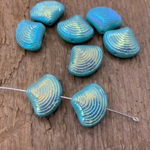 Puede incluir: Cuentas de concha azul iridiscente con una superficie texturizada y acanalada. Las cuentas son una mezcla de azul claro y dorado, con un brillo nacarado. Algunas cuentas están ensartadas en un alambre fino, otras sueltas. Las cuentas están sobre una superficie de madera.