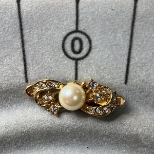 Puede incluir: Un cierre dorado con una perla central y cristales transparentes incrustados. El cierre tiene un diseño en forma de lazo, con la perla en el centro. Los cristales añaden brillo a la pieza.