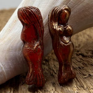 May include: Two small, red, metallic figurines of women with long hair. The figurines are standing upright and have a slightly distressed finish.