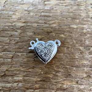 Puede incluir: Un cierre plateado en forma de corazón con un centro tachonado de pedrería. El cierre tiene un pequeño cierre de gancho y lazo. Se encuentra sobre una superficie de madera marrón texturizada.