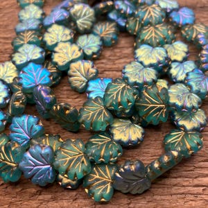 May include: A strand of iridescent teal glass beads in the shape of leaves. The beads have a gold metallic finish and are strung together on a clear thread.