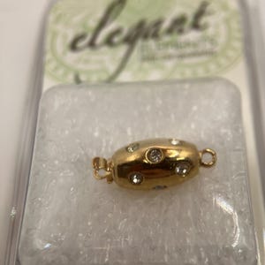 Puede incluir: Un cierre de joyería dorado con varios diamantes de imitación redondos y transparentes. El cierre tiene forma ovalada y tiene un pequeño lazo en cada extremo para sujetarlo a la joyería. El embalaje tiene la palabra "Elegant".
