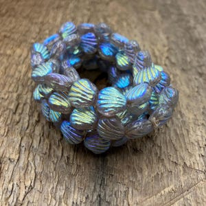 May include: A strand of iridescent glass beads in the shape of seashells. The beads are a light blue colour with a shimmery finish.