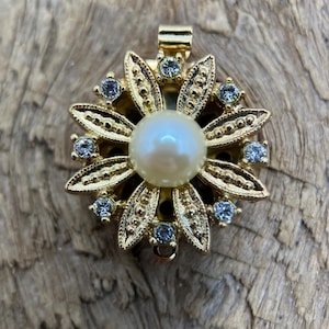 Puede incluir: Colgante dorado con diseño floral. Presenta una perla central rodeada de pétalos dorados y pequeñas gemas transparentes. El colgante tiene un lazo en la parte superior para una cadena.