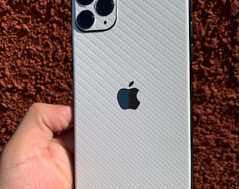 May include: A white iPhone with a textured, carbon fiber-like pattern on the back. The phone has a black camera module with three lenses and a flash. The Apple logo is visible. The phone is held in a hand.