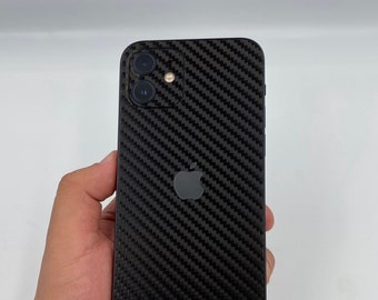 May include: Black carbon fiber textured phone case with the Apple logo. The case covers the back of the phone and has cutouts for the camera lenses. The phone is held in a hand against a white background.