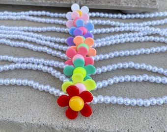 May include: A collection of white pearl necklaces with a vertical row of colorful flower-shaped beads. The flower beads are in various colors, including red, yellow, pink, green, orange, purple, and white. The pearls are small and round.