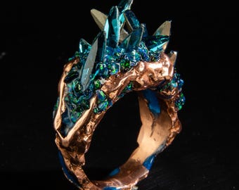 May include: A unique ring featuring a cluster of blue-green crystals atop a textured, copper-colored band. The ring has a dark background, highlighting the intricate details and vibrant colors of the gemstone and metalwork.