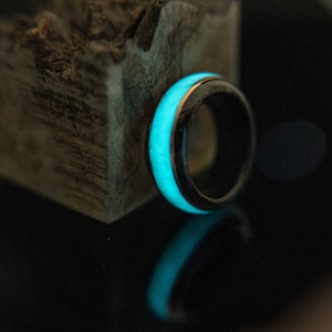 May include: A black ring with a bright blue glow-in-the-dark band. The ring is sitting on a piece of wood.