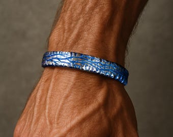 May include: A rigid bracelet featuring a textured surface with a metallic silver base and deep blue accents. The design consists of intricate, raised, branch-like patterns that wrap around the circumference of the band, creating a high-contrast look.