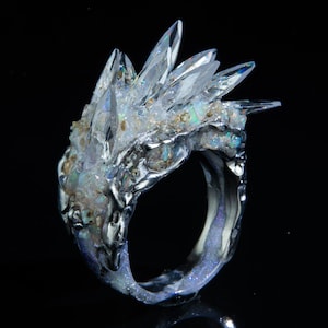 May include: A striking ring with a cluster of clear, pointed crystals on a textured, silver-coloured band. The ring has iridescent details and a dark background, emphasising the intricate design and craftsmanship of the jewellery.