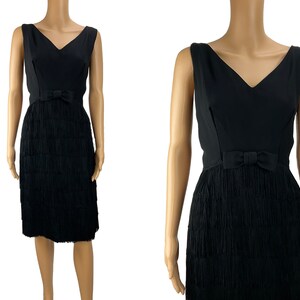 May include: A black sleeveless dress with a V-neckline and a bow at the waist. The skirt is made of fringe and is layered.
