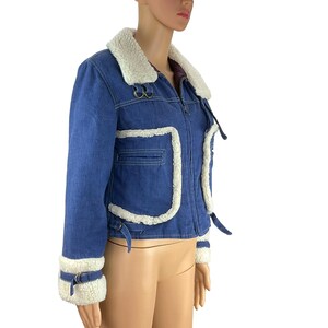 K&ouml;nnte beinhalten: Blaue Jeansjacke mit cremefarbenem Kunstfellkragen und -besatz. Die Jacke hat einen Rei&szlig;verschluss vorne, eine kleine Brusttasche und Schnallen an den &Auml;rmeln. Die Jacke ist ein verk&uuml;rzter Stil.