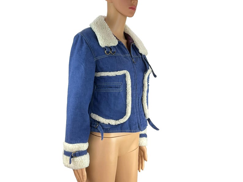 K&ouml;nnte beinhalten: Blaue Jeansjacke mit cremefarbenem Kunstfellkragen und -besatz. Die Jacke hat einen Rei&szlig;verschluss vorne, eine kleine Brusttasche und Schnallen an den &Auml;rmeln. Die Jacke ist ein verk&uuml;rzter Stil.