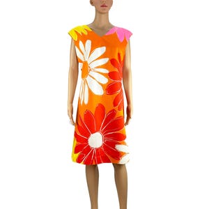 Può includere: Abito arancione, lunghezza al ginocchio, con grandi motivi floreali a margherita bianchi e rossi. L'abito ha uno scollo a V e maniche a cappuccio. L'abito è su un manichino.