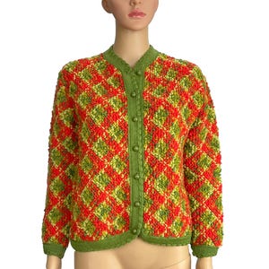 Può includere: Un cardigan vintage con bottoni e un motivo a rombi. Il maglione presenta una maglia strutturata nei toni del rosso, arancione e verde, con finiture e bottoni verdi. Il cardigan è esposto su un manichino.