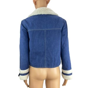 K&ouml;nnte beinhalten: Blaue Jeansjacke mit wei&szlig;em Kunstfellkragen und -b&uuml;ndchen. Die Jacke hat ein klassisches Design mit Knopfverschluss und eine verk&uuml;rzte Passform. Die Manschetten haben eine wei&szlig;e Kunstfellverbr&auml;mung.