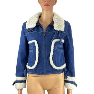 K&ouml;nnte beinhalten: Blaue Jeansjacke mit wei&szlig;em Kunstfellbesatz. Die Jacke hat einen Rei&szlig;verschluss, einen wei&szlig;en Kunstfellkragen und wei&szlig;e Kunstfellakzente an den Taschen und Manschetten. Die Jacke hat einen klassischen, Vintage-Stil.