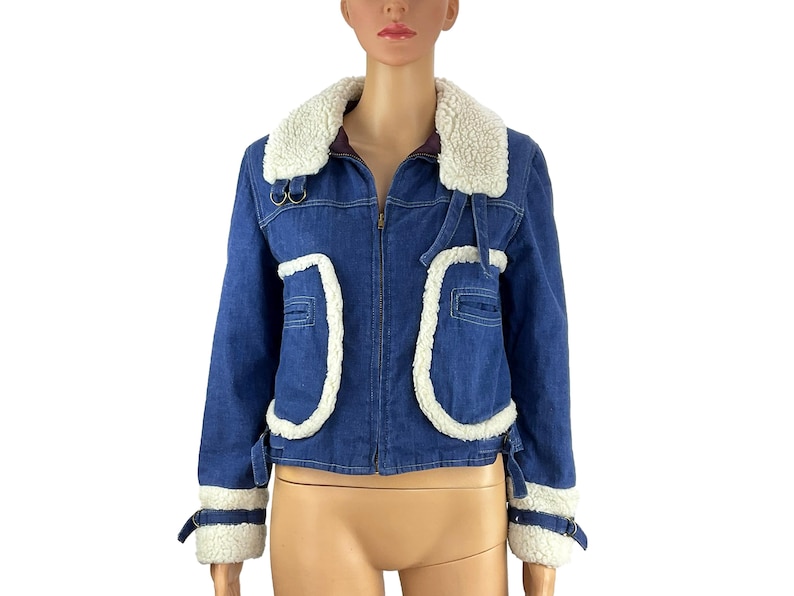 K&ouml;nnte beinhalten: Blaue Jeansjacke mit wei&szlig;em Kunstfellbesatz. Die Jacke hat einen Rei&szlig;verschluss, einen wei&szlig;en Kunstfellkragen und wei&szlig;e Kunstfellakzente an den Taschen und Manschetten. Die Jacke hat einen klassischen, Vintage-Stil.