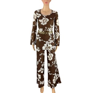 May include: A brown jumpsuit with a white floral pattern. The long-sleeved top has a scoop neckline and the pants are flared. A gold-colored belt with circular links cinches the waist. The jumpsuit is a vintage style.