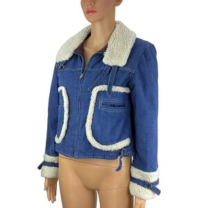 K&ouml;nnte beinhalten: Blaue Jeansjacke mit wei&szlig;em Kunstfellkragen und -besatz. Die Jacke hat einen Rei&szlig;verschluss, Taschen und dekorative Riemen an den &Auml;rmeln. Die Jacke wird auf einer Schaufensterpuppe pr&auml;sentiert.