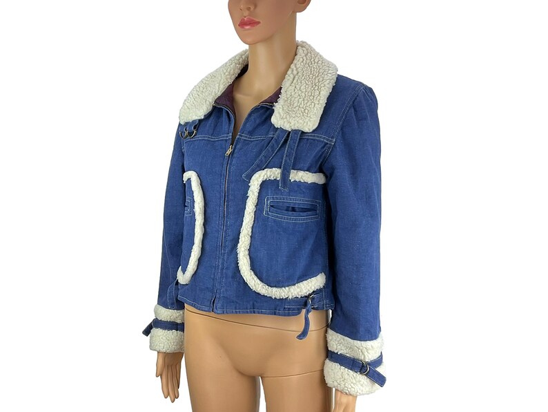 K&ouml;nnte beinhalten: Blaue Jeansjacke mit wei&szlig;em Kunstfellkragen und -besatz. Die Jacke hat einen Rei&szlig;verschluss, Taschen und dekorative Riemen an den &Auml;rmeln. Die Jacke wird auf einer Schaufensterpuppe pr&auml;sentiert.