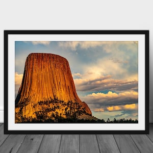 May include: A large, orange-brown rock formation, known as Devils Tower, rises dramatically against a backdrop of a blue sky with white clouds. The rock face is textured and appears to be made of many layers. The sky is bright and the clouds are fluffy.