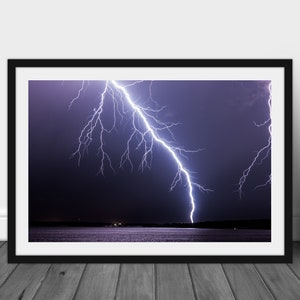 May include: A dramatic photograph of a lightning strike over a dark body of water. The lightning bolt is white and branches out in a jagged pattern against a dark purple sky.