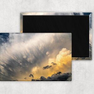 Puede incluir: Imán rectangular con una escena de cielo dramático con nubes arremolinadas en tonos de gris, blanco y amarillo. El imán tiene un respaldo negro y un borde plateado. Un segundo imán es parcialmente visible, mostrando el mismo diseño.