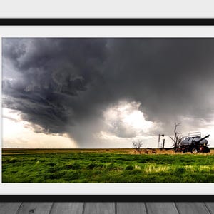 May include: A dark, stormy sky hangs over a grassy field with a lone, weathered truck parked in the distance. A windmill stands tall in the background, silhouetted against the ominous clouds.