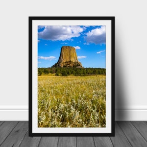 Könnte beinhalten: Eine große, gelbe Felsformation, bekannt als Devils Tower, ragt aus einem grasbewachsenen Feld empor. Der Himmel ist blau mit weißen Wolken.