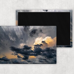 Puede incluir: Imán rectangular que representa un cielo dramático con nubes oscuras y arremolinadas y una explosión de luz dorada. El imán tiene un borde negro y un acabado brillante, mostrando los colores vibrantes del atardecer o amanecer.