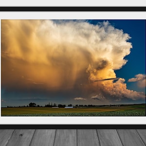 May include: A dramatic, golden-hued storm cloud hangs low over a green field, with a few trees and a small building in the distance.