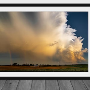 May include: A dramatic, golden-hued sky with a large, white cumulus cloud formation. A faint rainbow is visible in the distance, and a green field stretches out in the foreground.