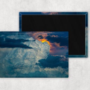 Puede incluir: Imán rectangular con un dramático paisaje de nubes en tonos azules, blancos y naranjas. La imagen muestra una gran formación de nubes esponjosas con una vibrante puesta de sol o amanecer en el fondo. El imán tiene un borde azul oscuro.