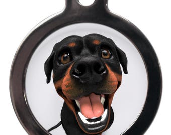 May include: A round, silver-colored pet tag featuring a smiling Rottweiler dog. The dog has black fur with brown markings and a pink tongue. The tag has a hole at the top for attaching to a collar.