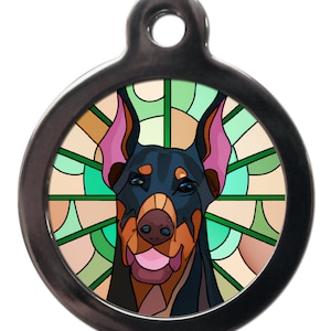 May include: A round, silver dog tag with a stained glass design featuring a Doberman Pinscher. The dog's head is centred in the design, with a green, blue, and brown stained glass background.